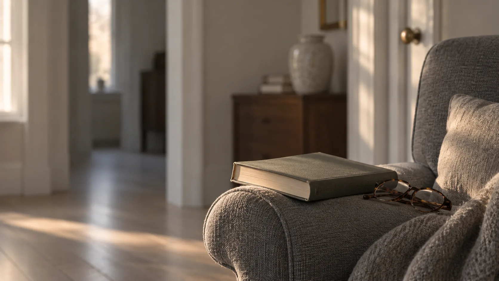 An open book laid face down on the rolled arm of a grey upholstered chair with a pair of tortoiseshell reading glasses folded beside it, warm light across the fabric and a dim room behind.